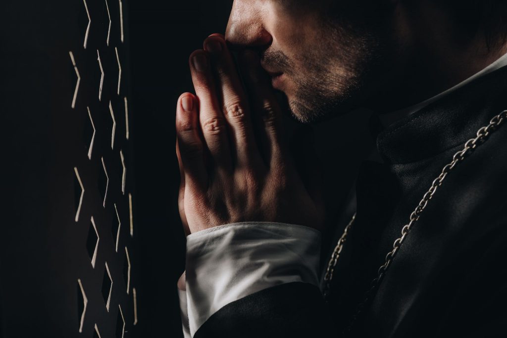 young catholic priest praying with closed eyes near confessional grille