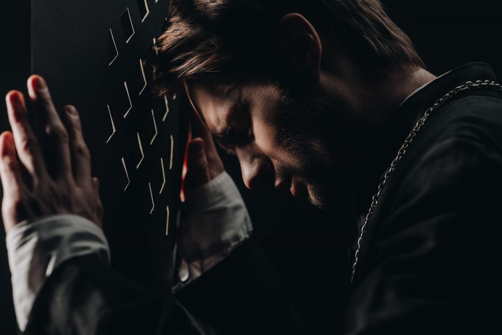 young tense catholic priest leaning on confessional grille in dark