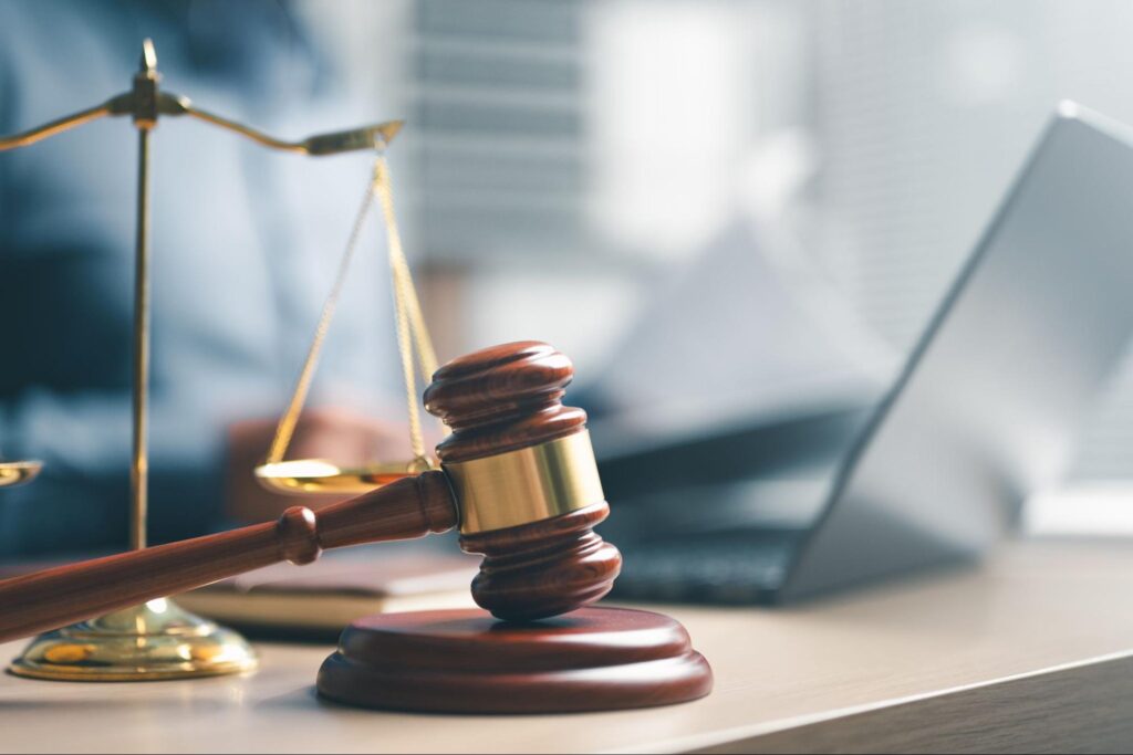 A wooden gavel and justice scale placed on a desk with a laptop in the background.